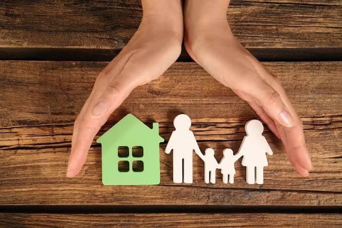 Hands protecting a family and home made of wooden figures on a rustic background.